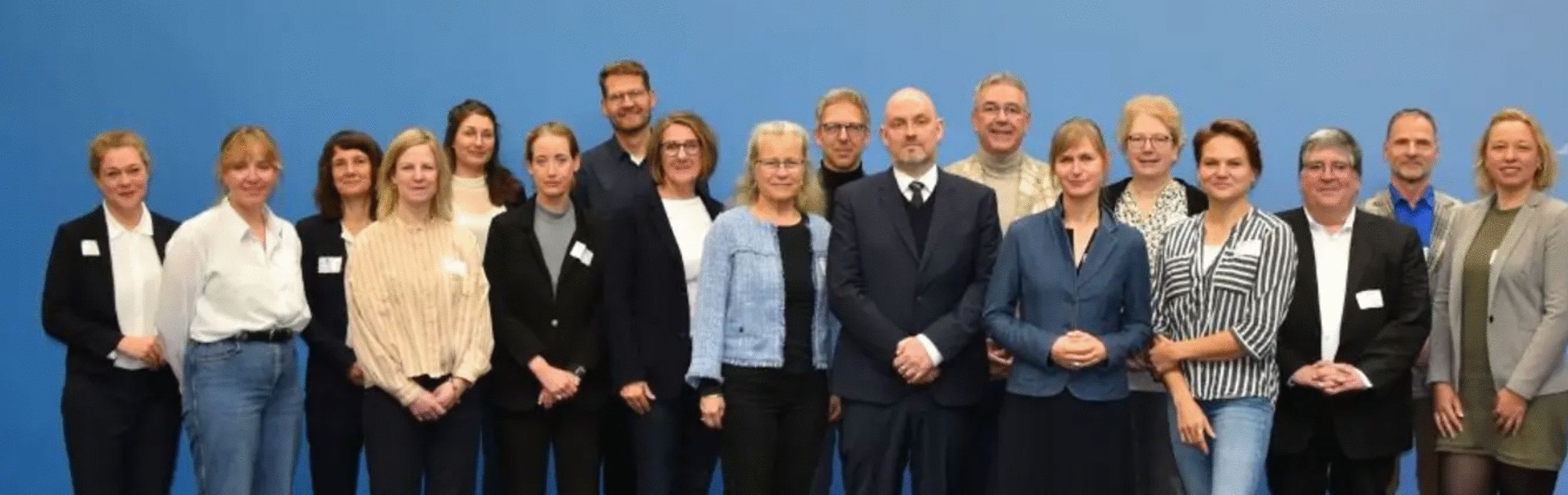 Eine große Gruppe Menschen steht vor einer hellblauen Wand und blickt in die Kamera.