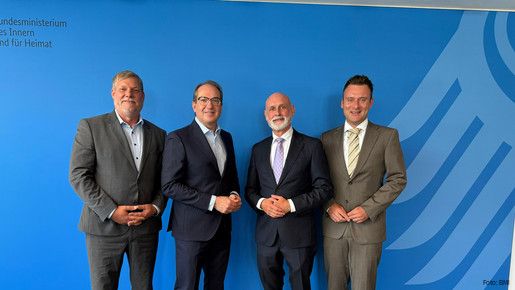 Vier Männer in Anzügen stehen vor einer blauen Wand und blicken in die Kamera