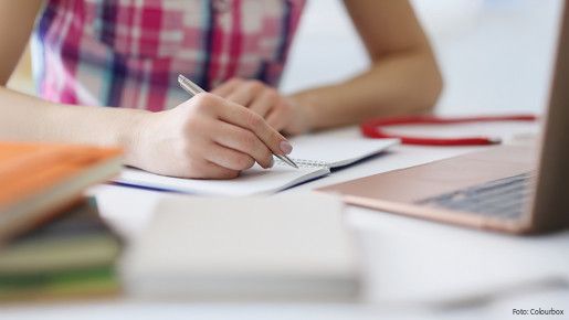 Das Foto zeigt die Hand einer Person, die mit einem Stift auf einem Block schreibt. Rechts im Bild steht ein aufgeklapper Laptop.