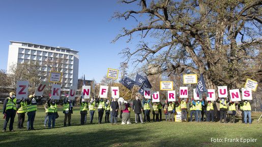 Eine Gruppe von Menschen steht auf einer Grünfläche und protestiert. Sie halten Schilder hoch die zusammen gesetzt "Zukunft nur mit uns" darstellen 