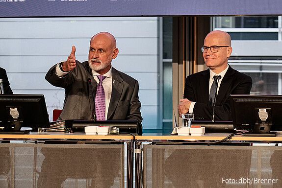 Zwei Männer in Anzügen sitzen an einem Tisch uns sprechen in ein Mikrofon